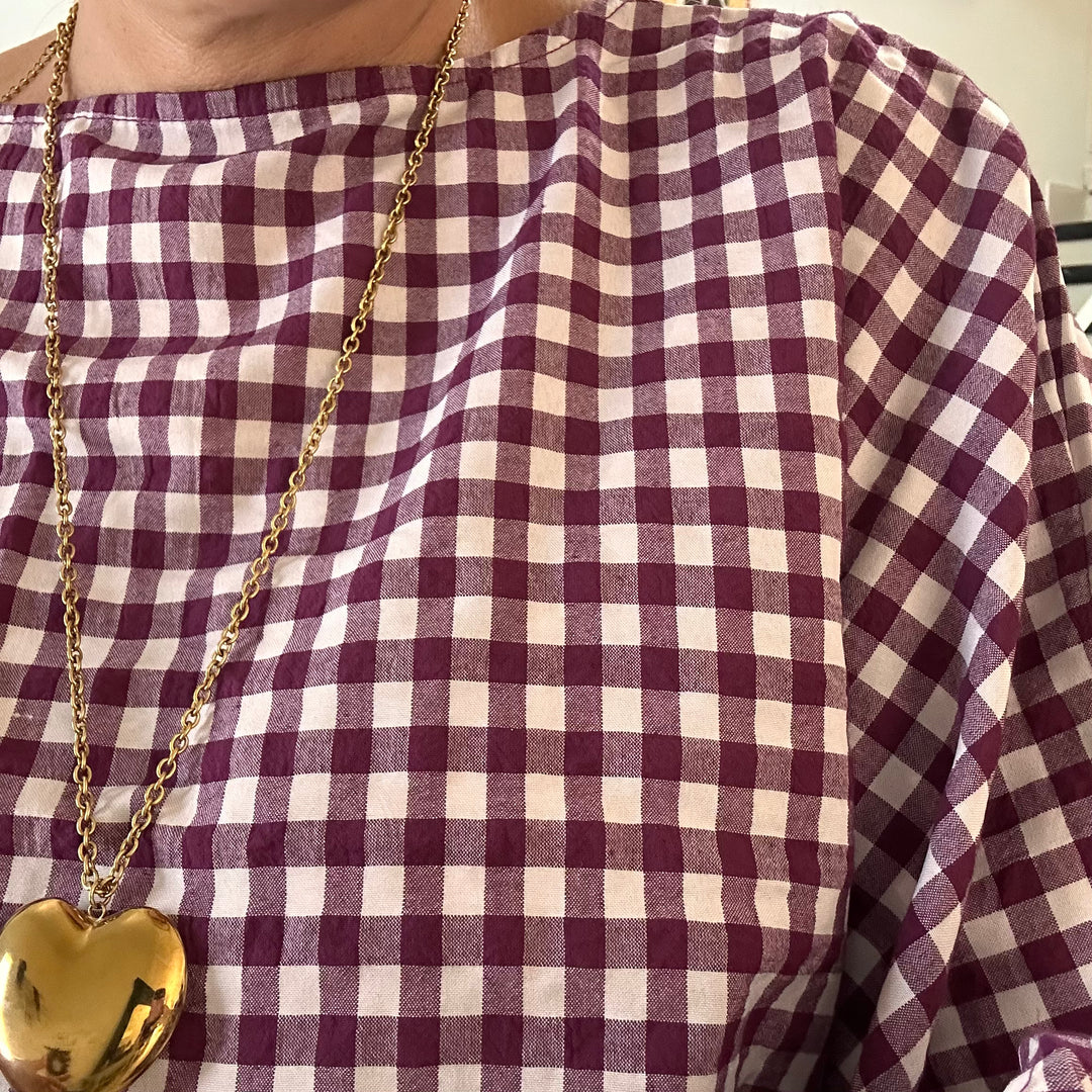A close-up of a person wearing a burgundy and cream gingham check top, accessorized with a gold chain necklace featuring a polished gold heart pendant.