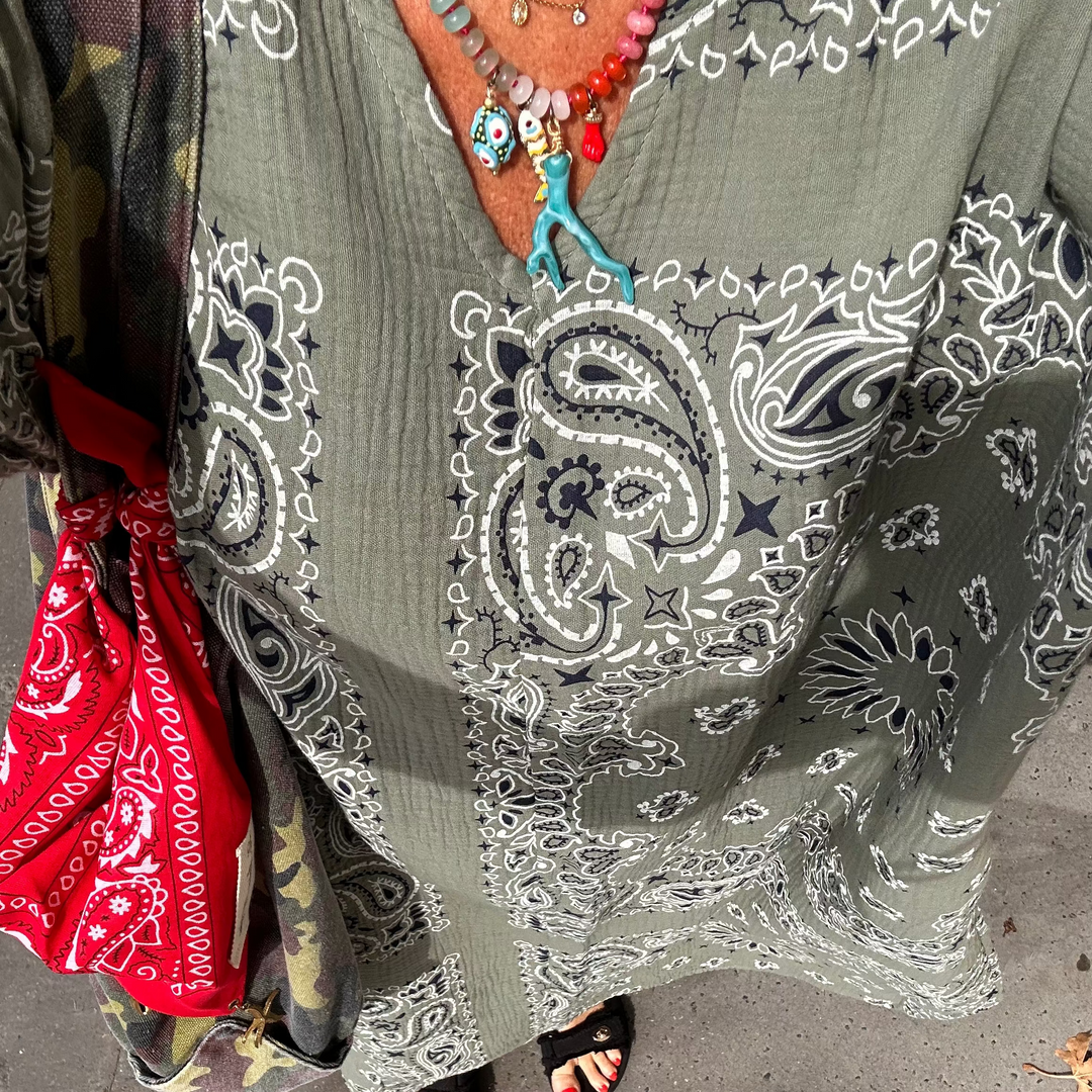 A person wearing a loose kaki green kaftan cotton gauze dress with a white and navy paisley bandana print, styled with a red bandana bag, colorful beaded necklaces