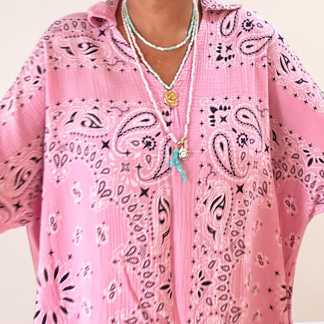 A pink bandana print kaftan top with black paisley patterns, worn by a model layered with bohemian necklaces.